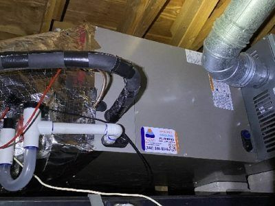 HVAC unit in an attic. Silver metal box with white pipes and insulated tubing, surrounded by wood beams.