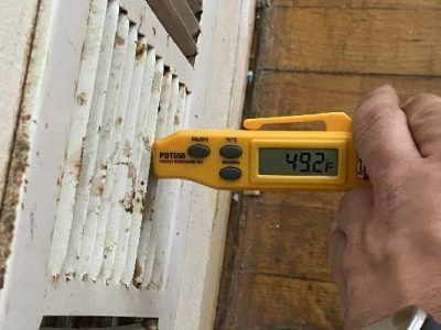 A person holding a yellow digital thermometer measuring 49.2°F on a white vent.