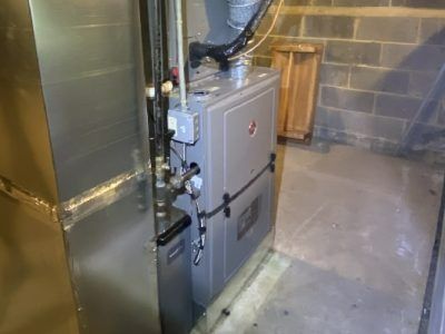 Furnace in a basement; gray metal unit with connected ductwork and pipes, placed near cinder block wall.