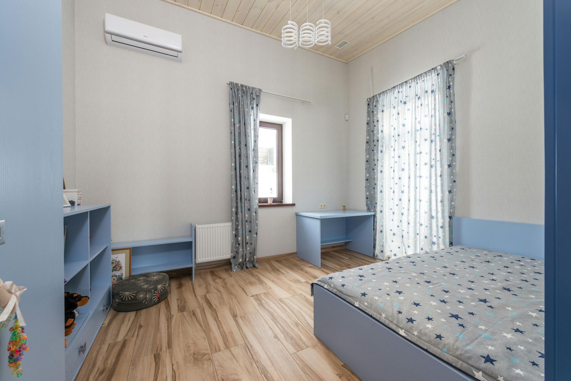 Blue-themed bedroom with wood floors. A bed, desk, and shelving sit near a window with starry curtains.