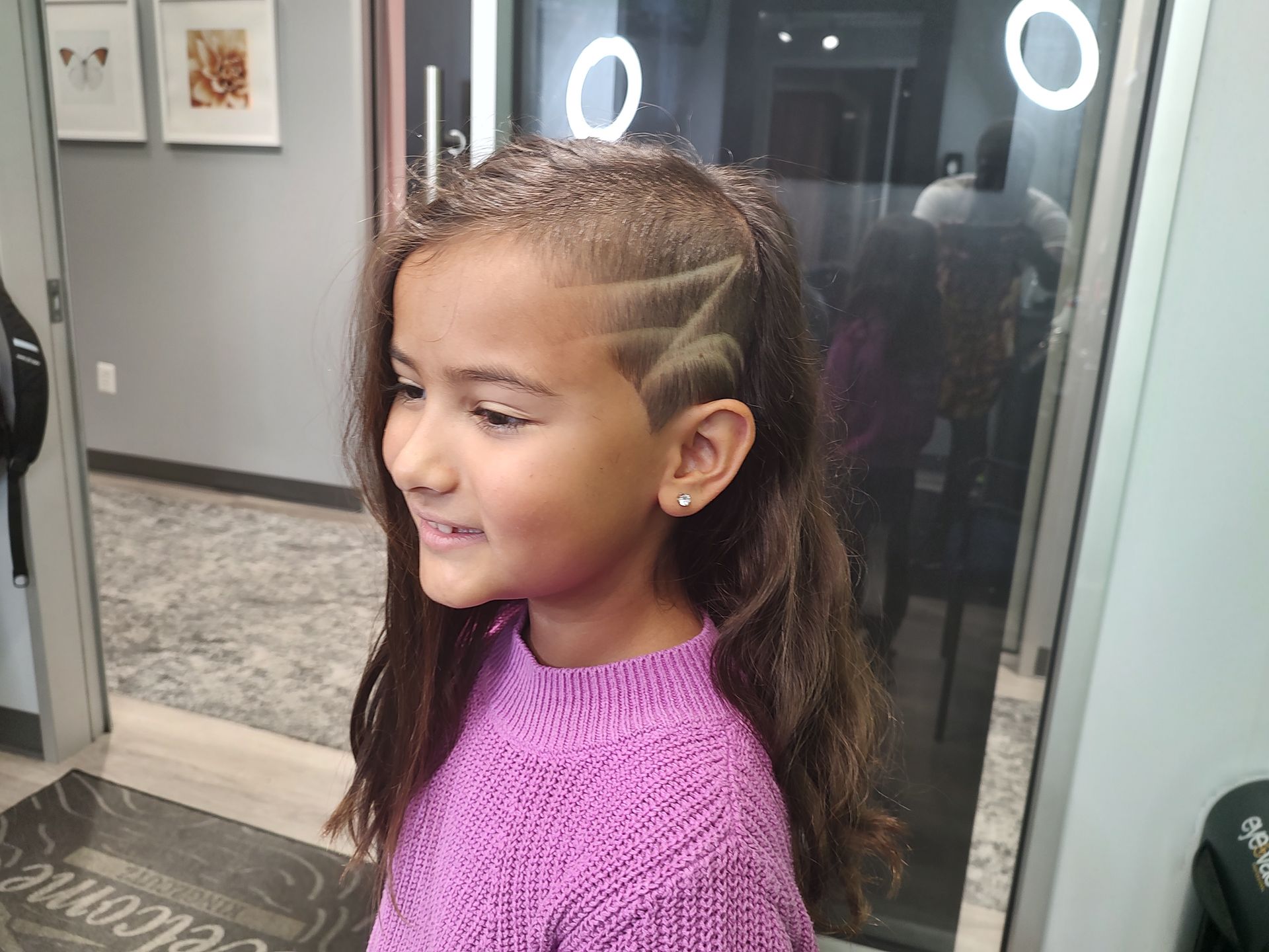 A little girl with a shaved design in her hair is wearing a purple sweater.