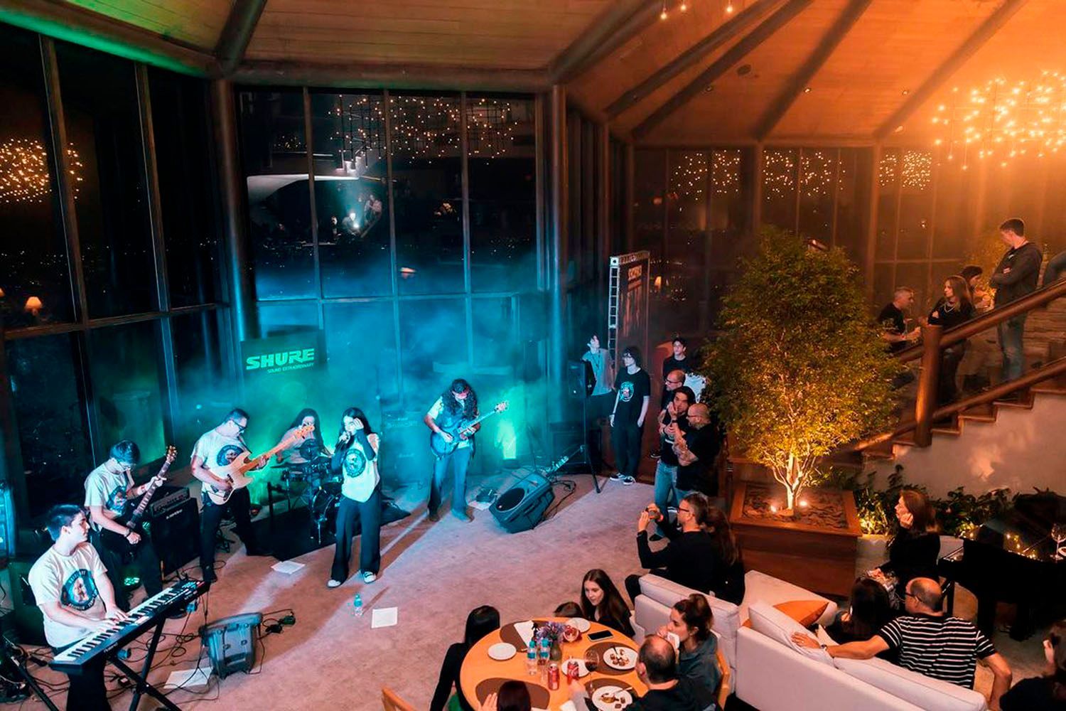 Um grupo de pessoas está sentado em mesas em uma sala grande assistindo a uma banda se apresentar no palco.