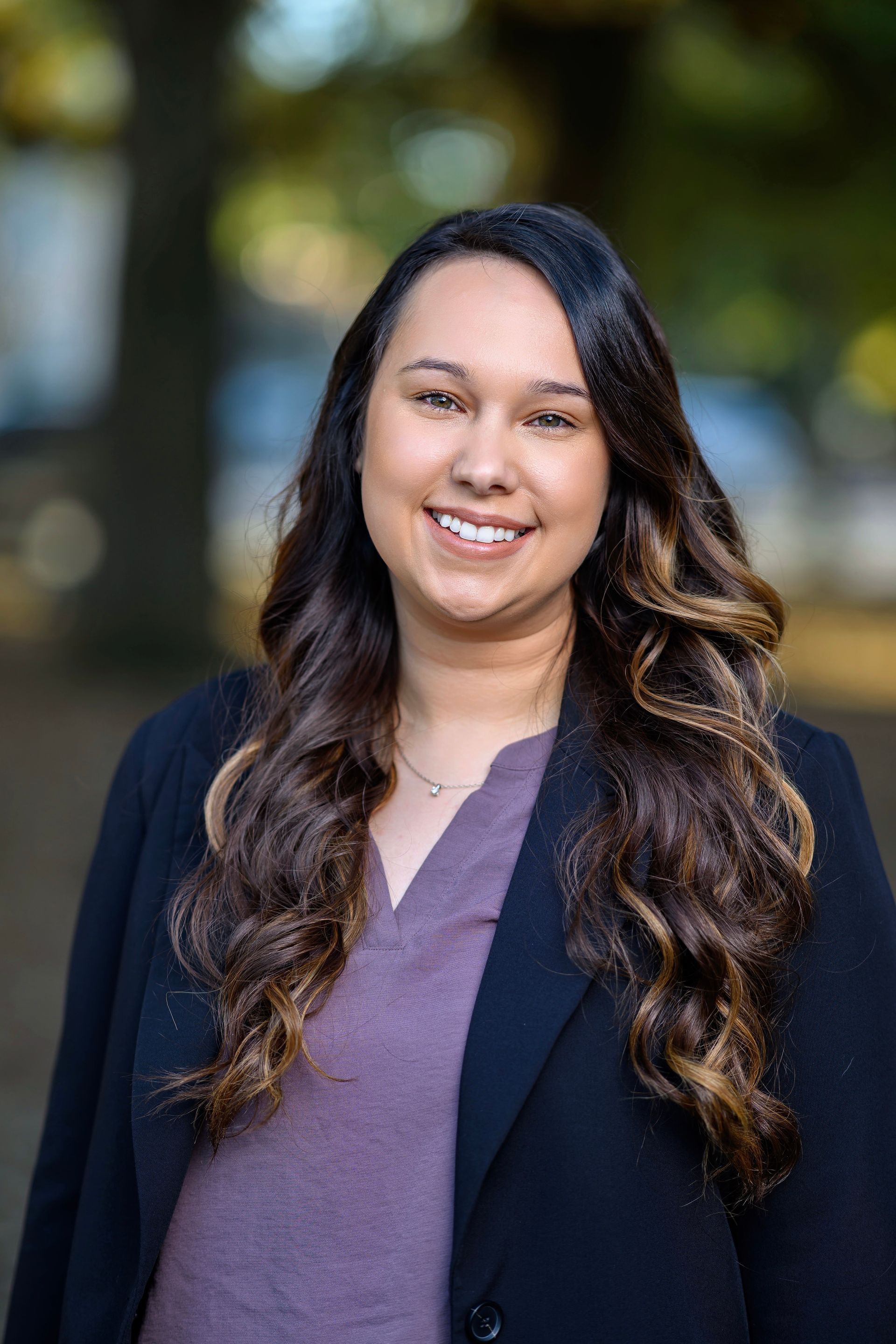 Woman with long wavy brown hair smiles outdoors, wearing a blazer over a purple top.