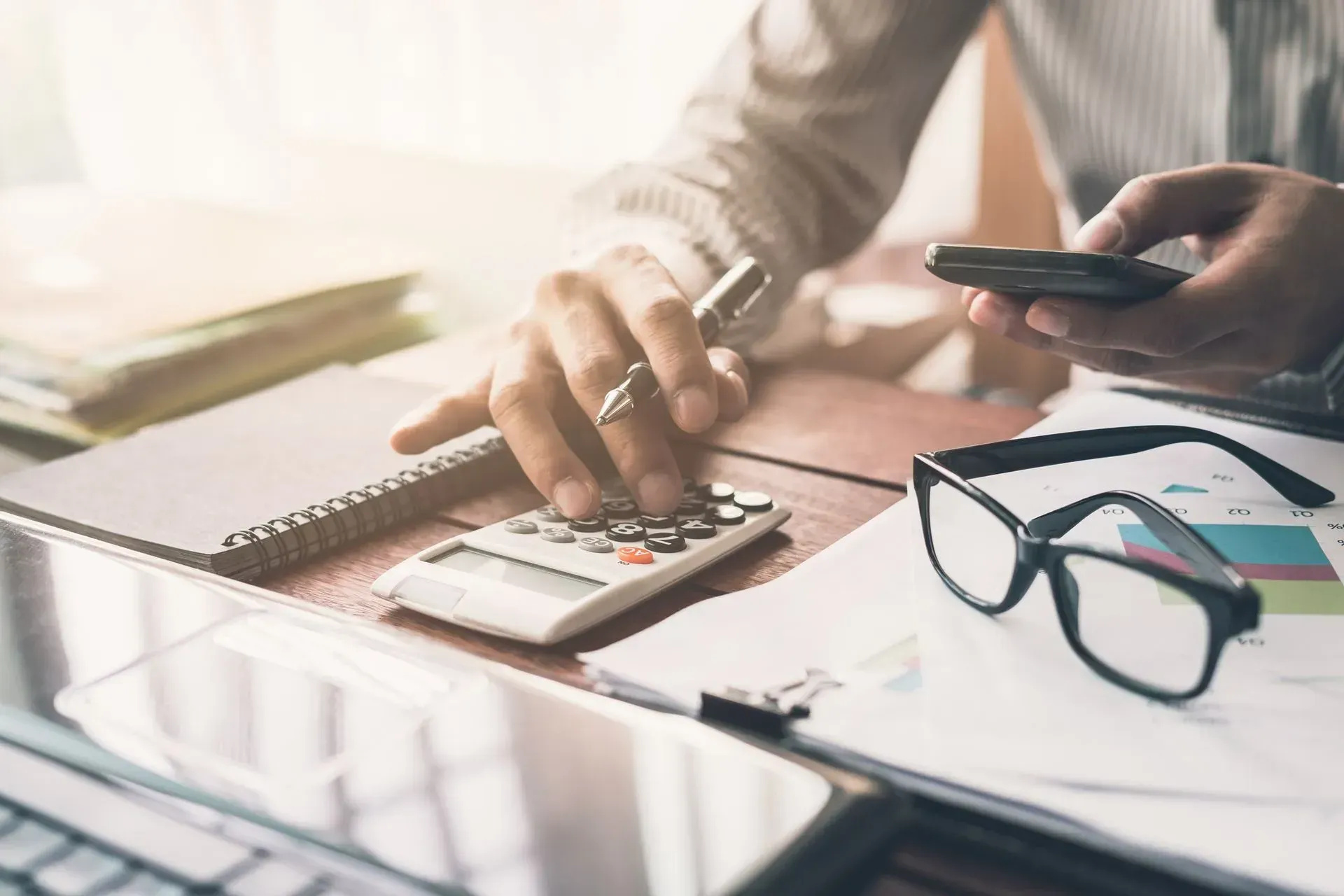 Hands using a calculator at a desk with glasses and a phone, sunlight on paperwork.