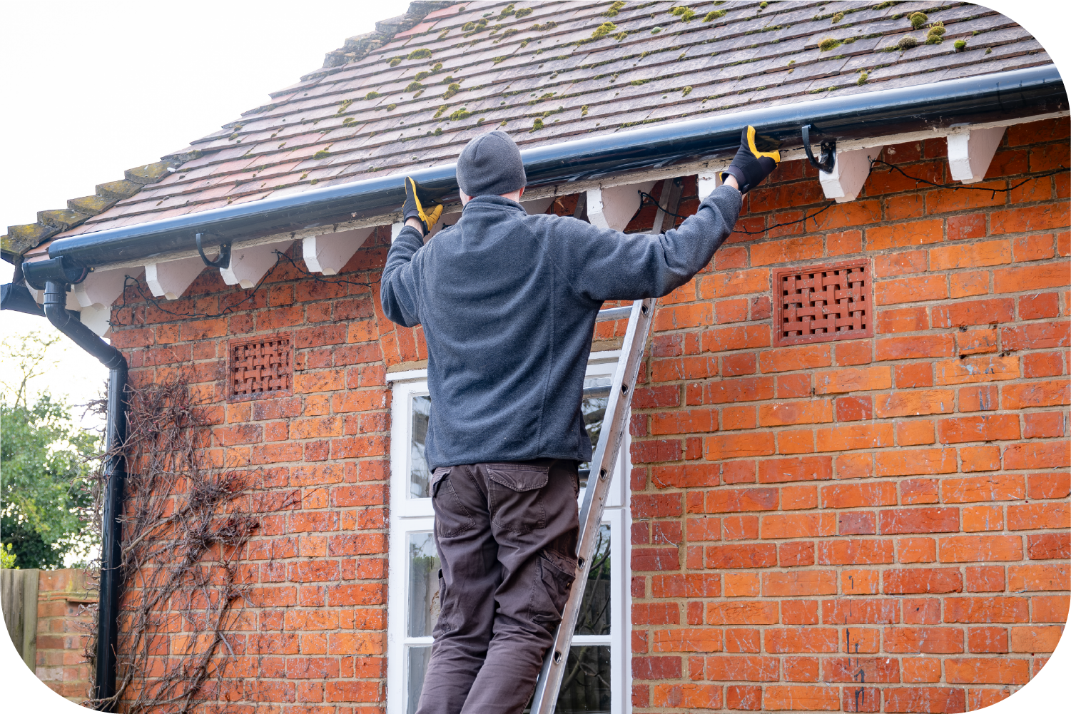 A man is standing on a ladder fixing a gutter on a brick building.