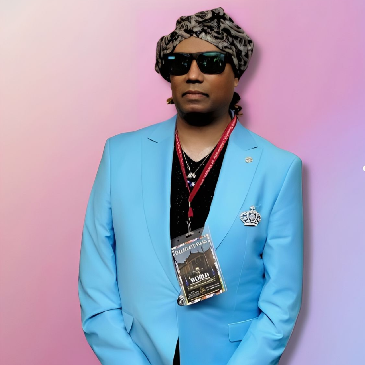 Man wearing sunglasses and turban, light blue suit. He has a lanyard and is in front of a colorful background.