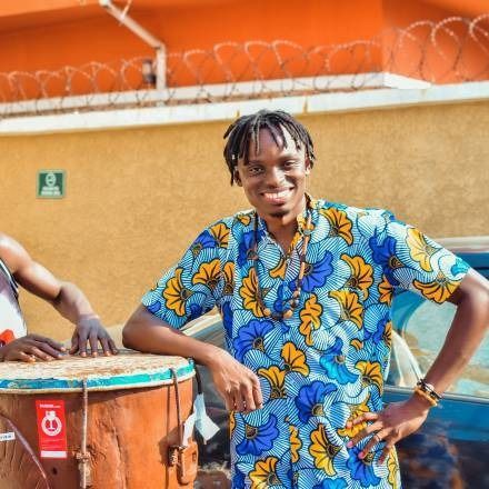 Man in blue patterned shirt smiles next to a drum. Leaning on drum, orange wall.