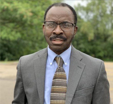 Man in glasses and suit, looking at the camera; outdoor setting with green background.