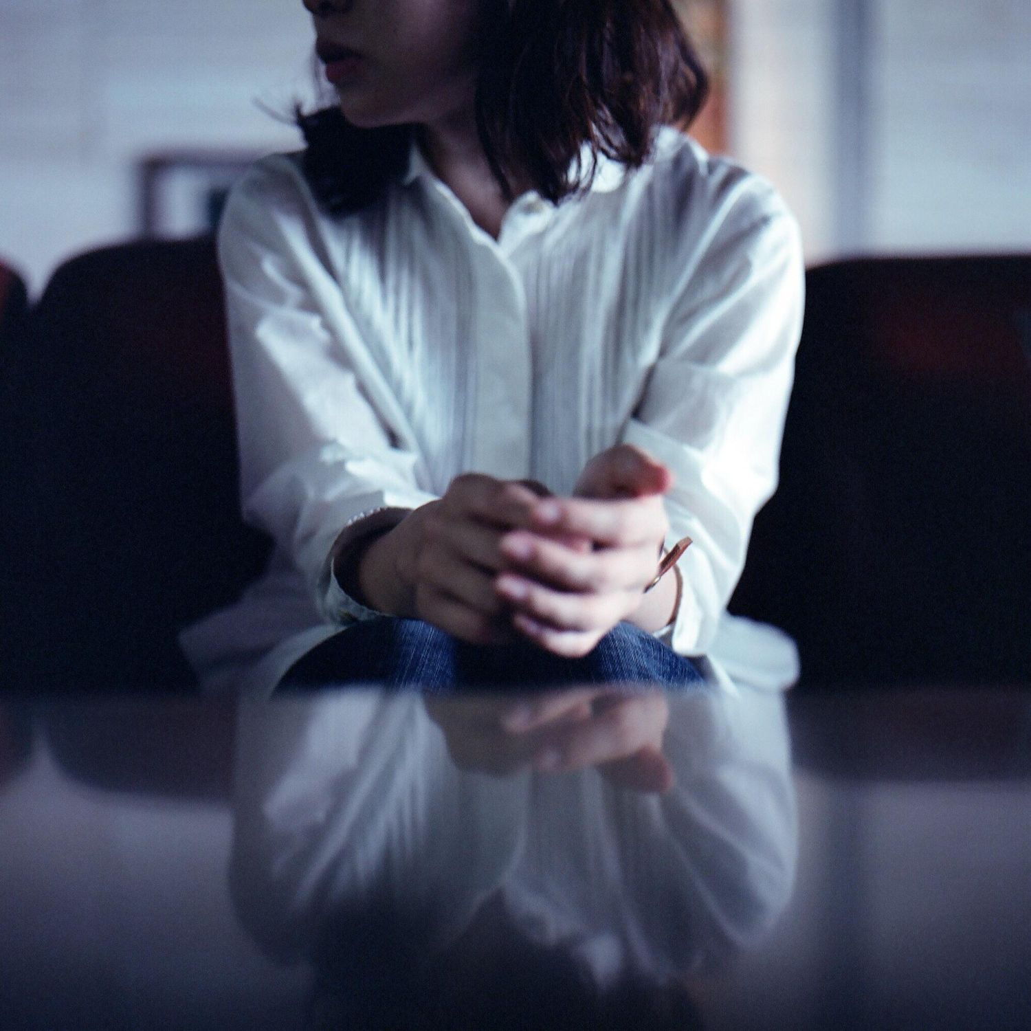 Woman in white shirt sits on a sofa, hands clasped. Reflection visible on a table.
