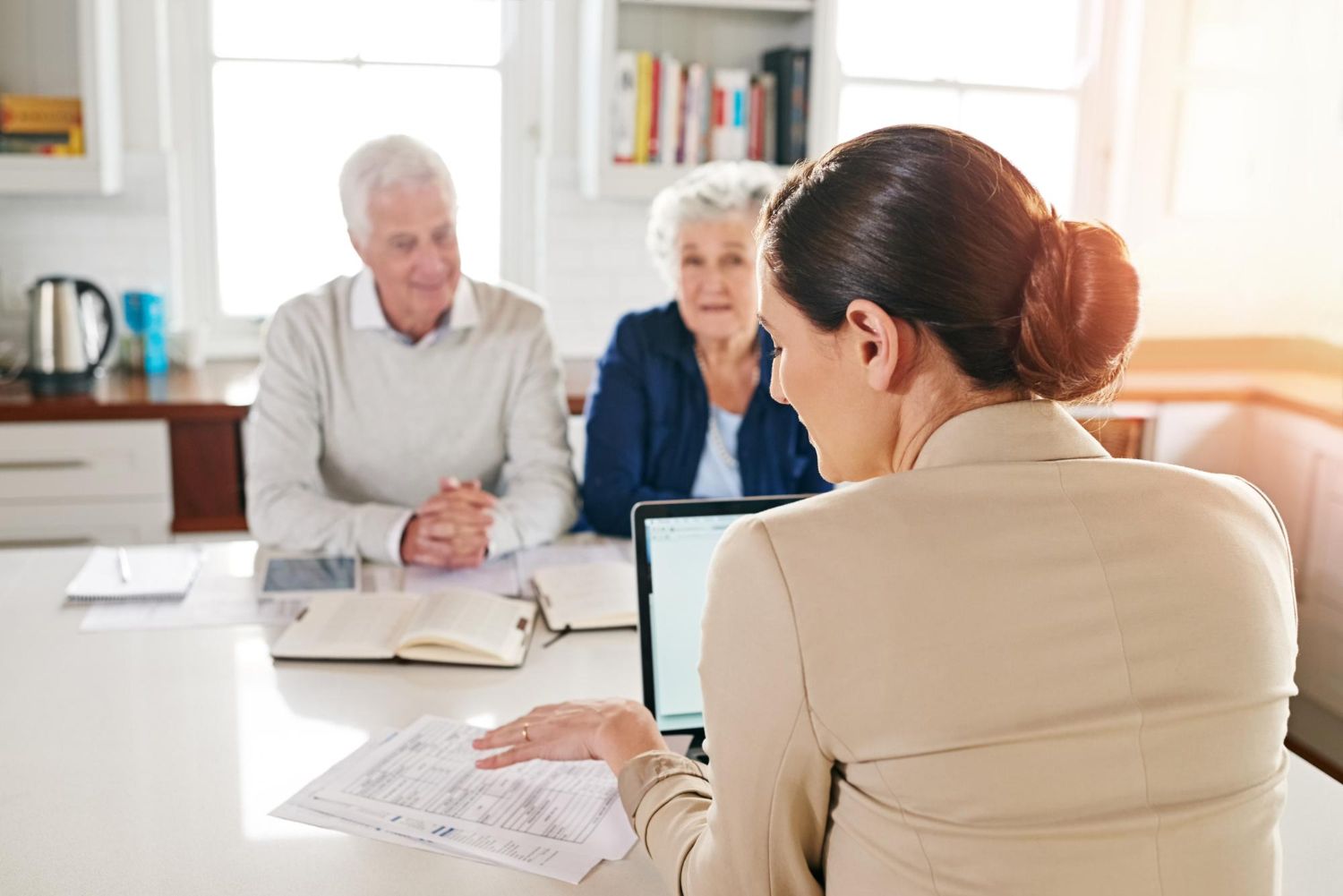 Woman showing papers to older couple sitting at a table with laptop in a bright room.