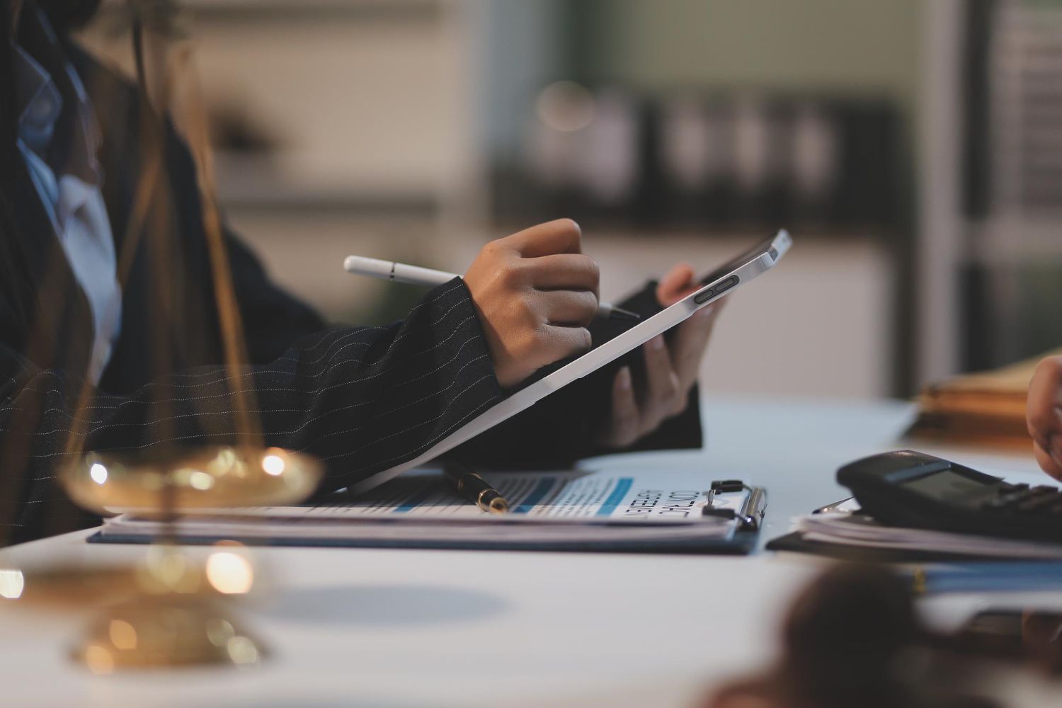 Person in a blazer using a tablet and pen over documents on a desk with scales of justice in the foreground.