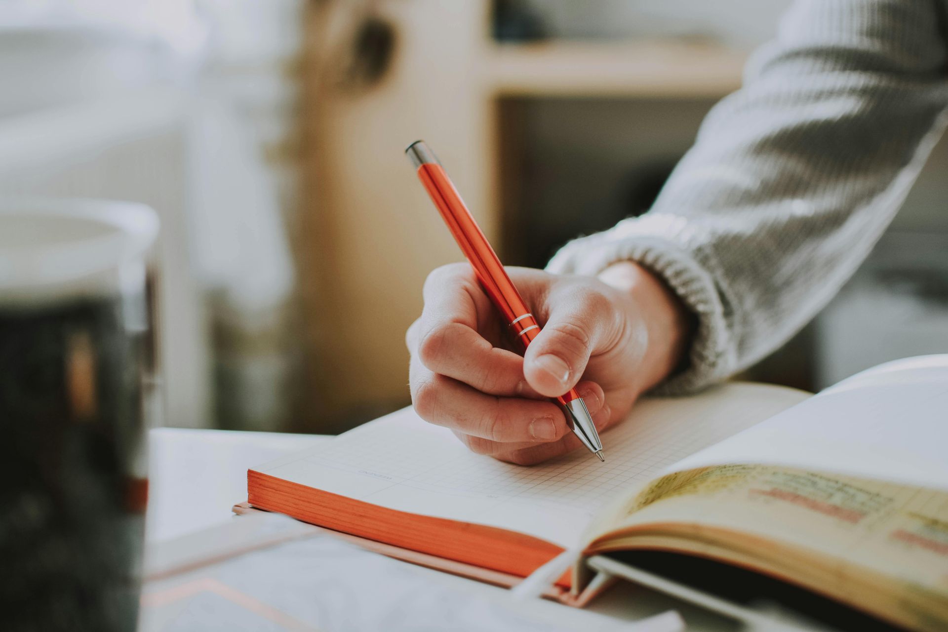 Person's hand writing with a red pen in an open notebook with a red bookmark on a desk.