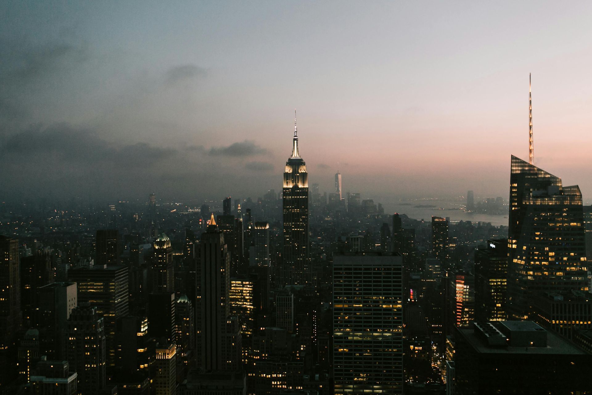 New York City skyline at dusk, lit buildings against a cloudy, pink-tinged sky.