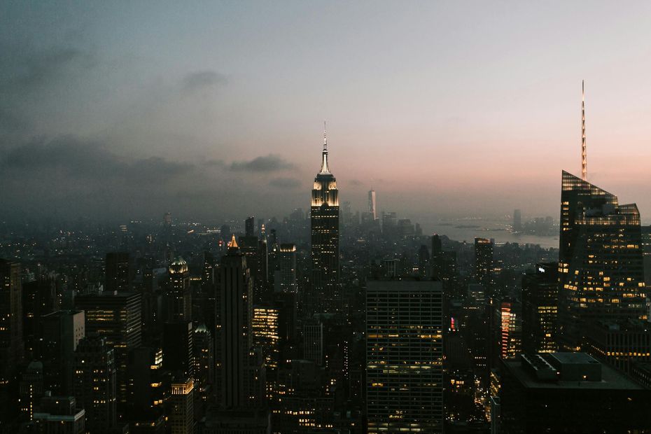New York City skyline at dusk, lit buildings against a cloudy, pink-tinged sky.