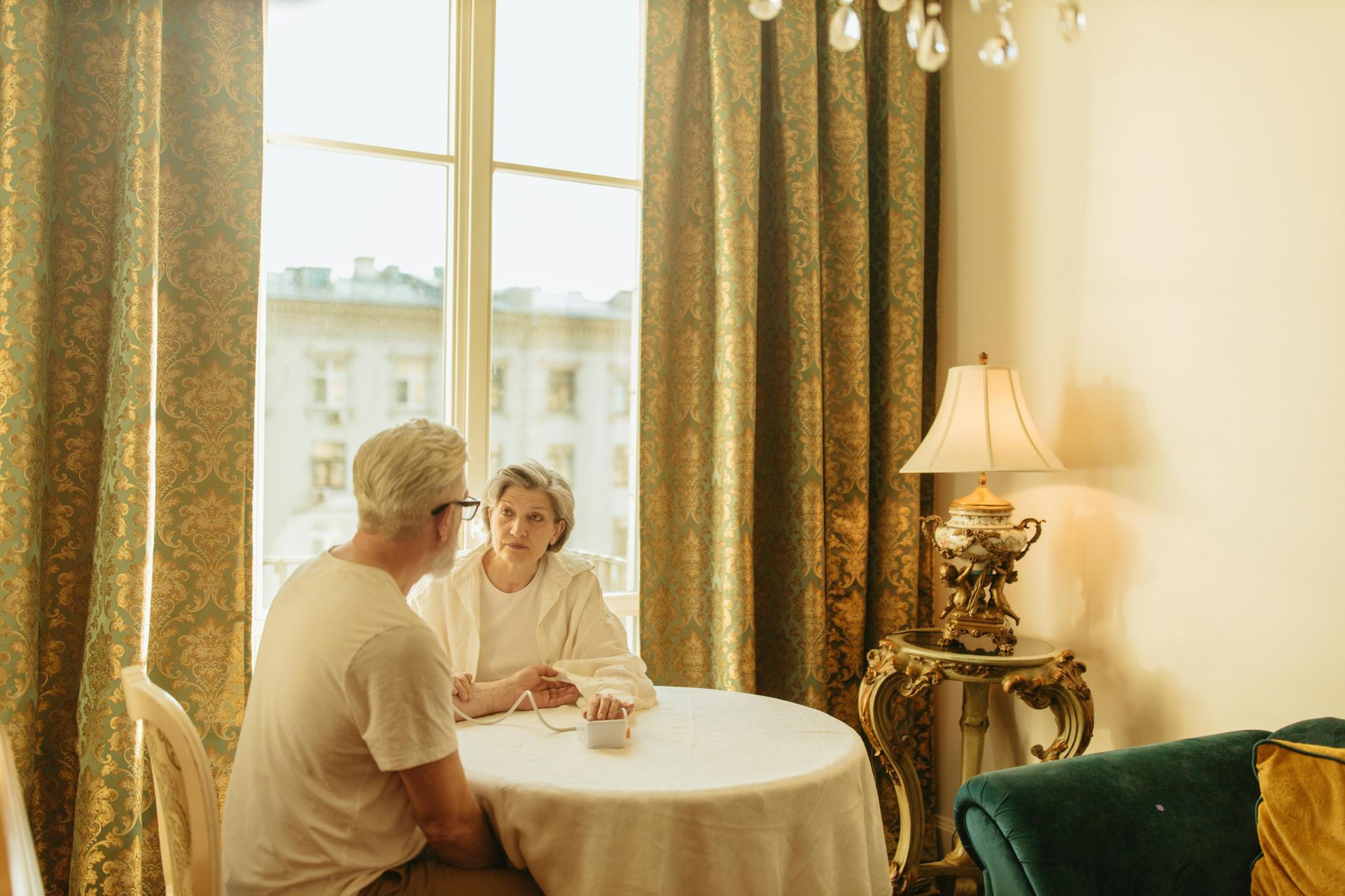An older couple sits at a round table by a window, talking. Warm lighting, ornate decor.