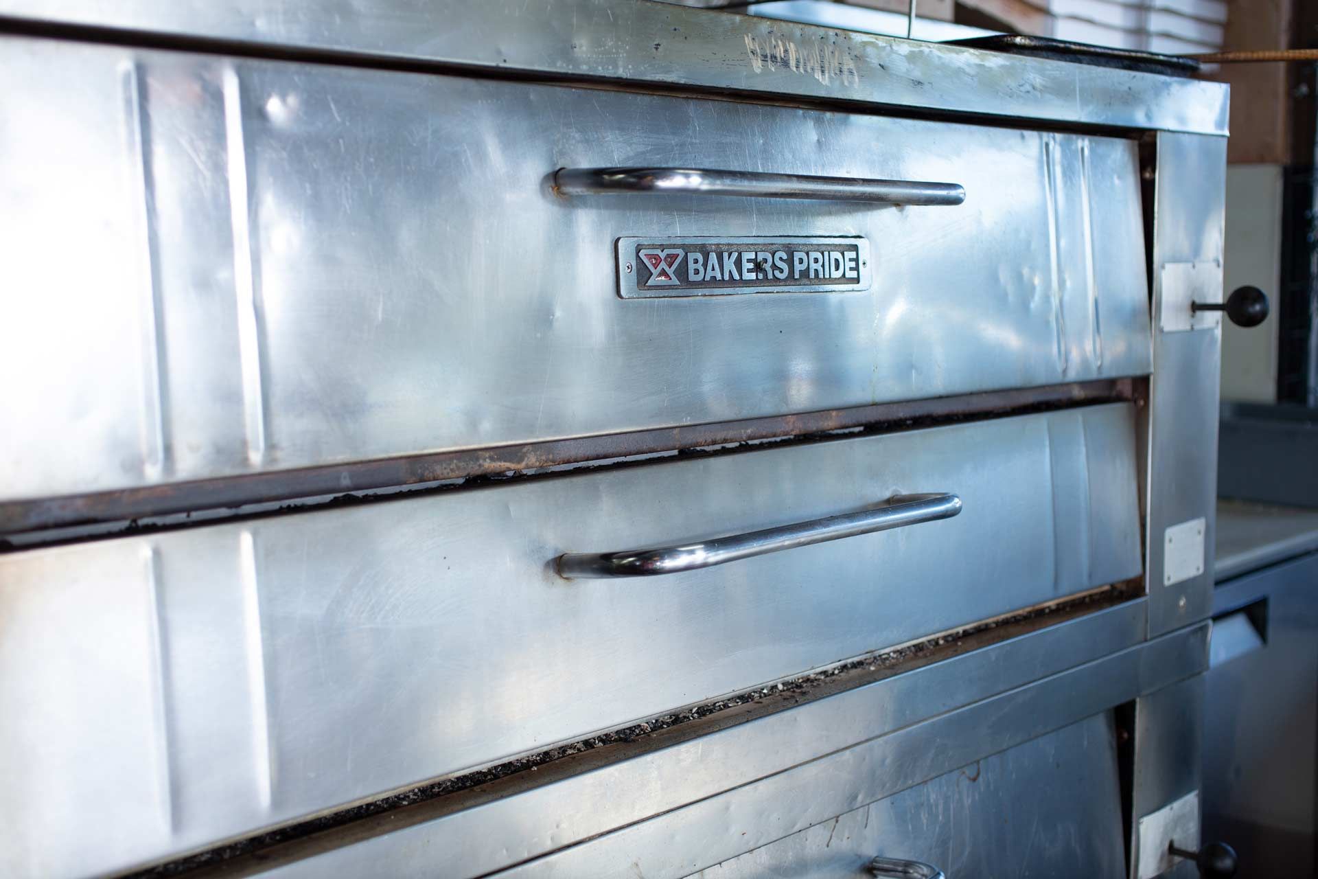 Man in blue overalls inspects a commercial oven in a kitchen. Another person is in the background.