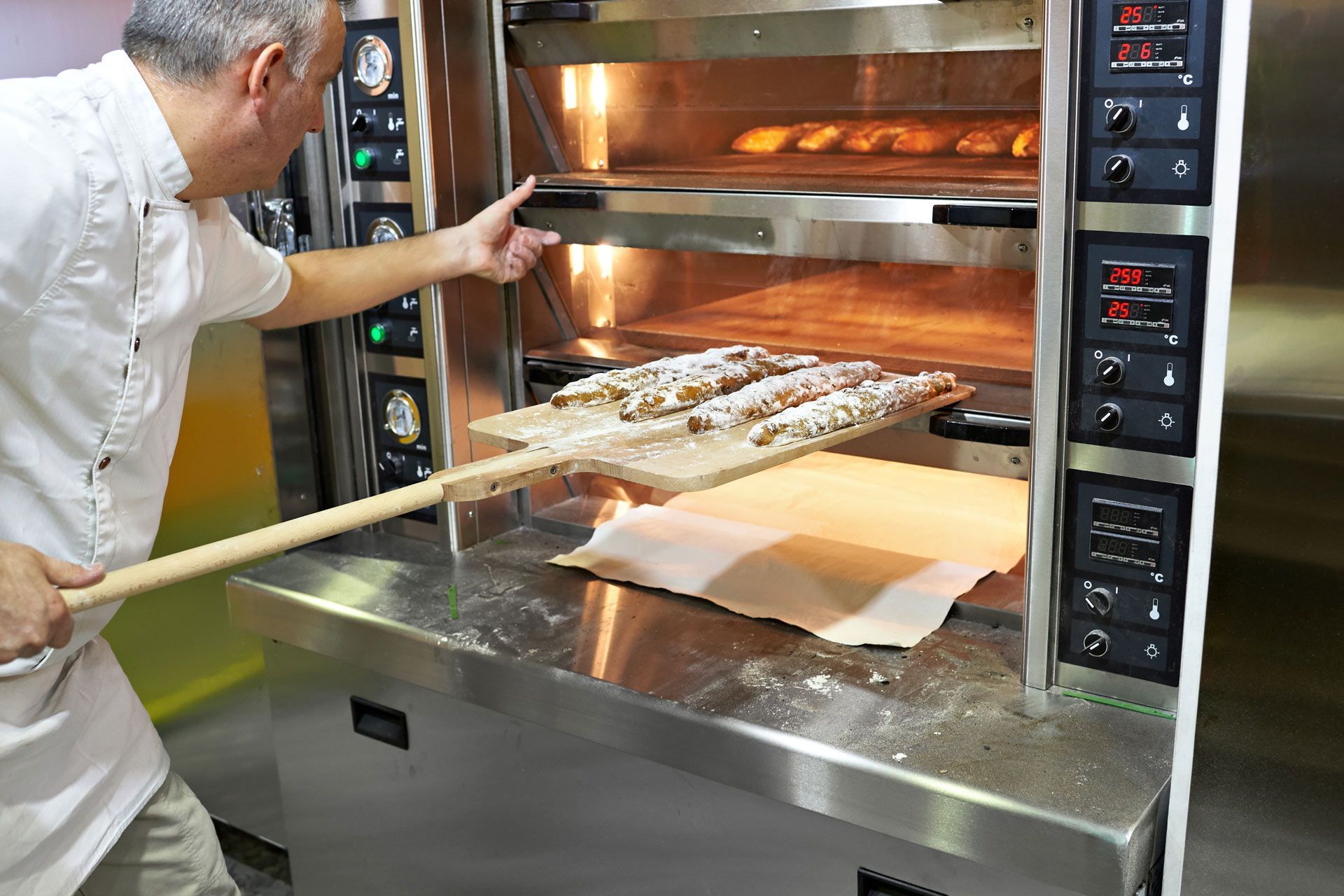 Baker placing loaves of bread into a commercial oven with a peel. Stainless steel, warm interior.