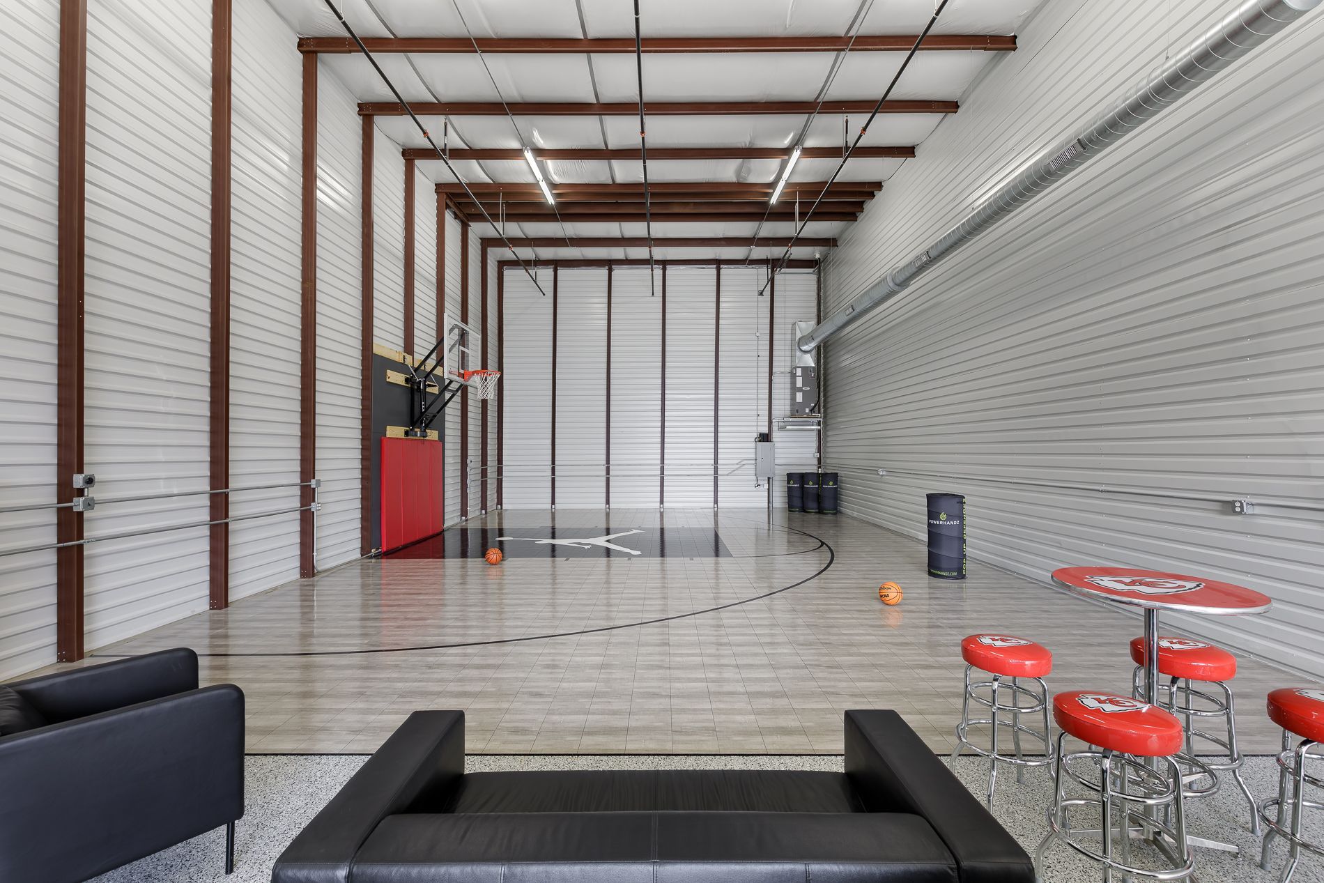 Indoor basketball court with red bar stools, black couches, and hoop; gray walls.