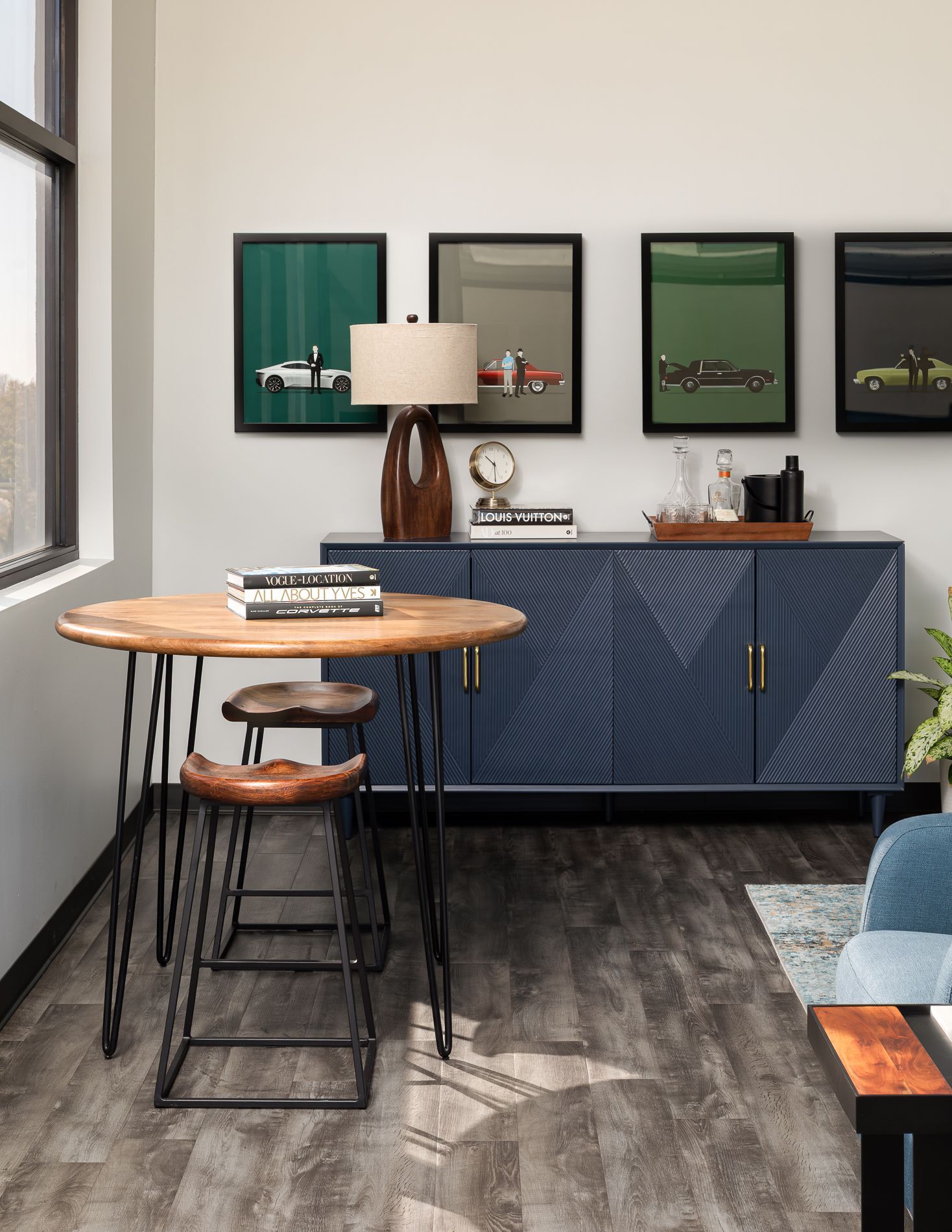 A modern dining area with a round wooden table, bar stools, blue cabinet, and framed car prints.