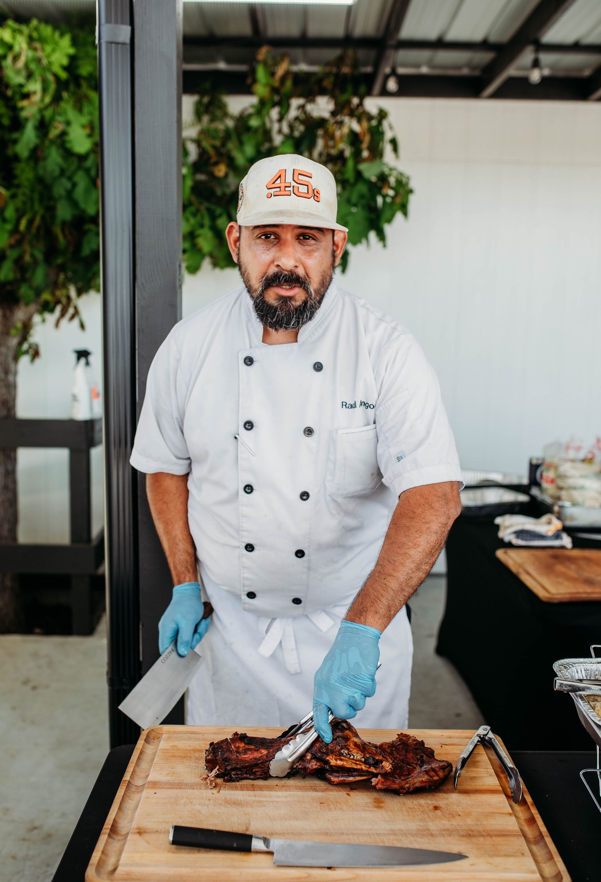 A chef's hat and blue gloves cuts a piece of meat on a wooden during an event