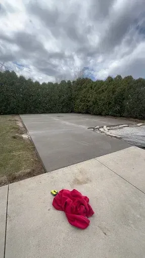 Newly poured concrete slab with a red cloth lying on a lighter concrete patio, backed by dense green trees under a cloudy sky.