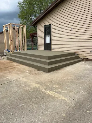 Concrete steps lead to a door on a tan house; a wooden frame stands to the left.