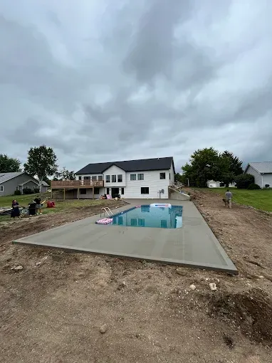 A new concrete pool deck with a pool in front of a two-story white house, on a cloudy day.