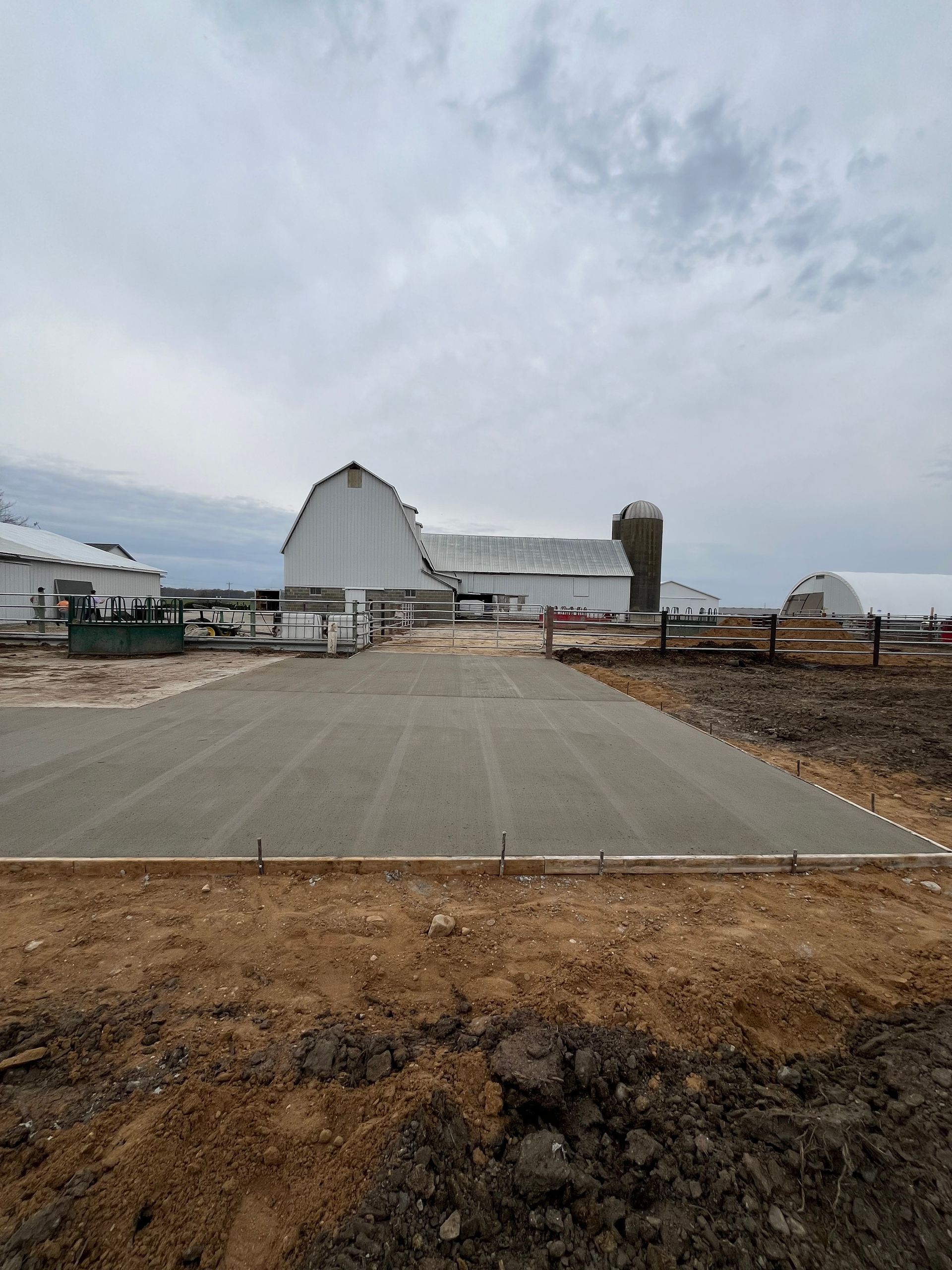 Newly poured concrete pad in front of a white barn with a silo, under an overcast sky.