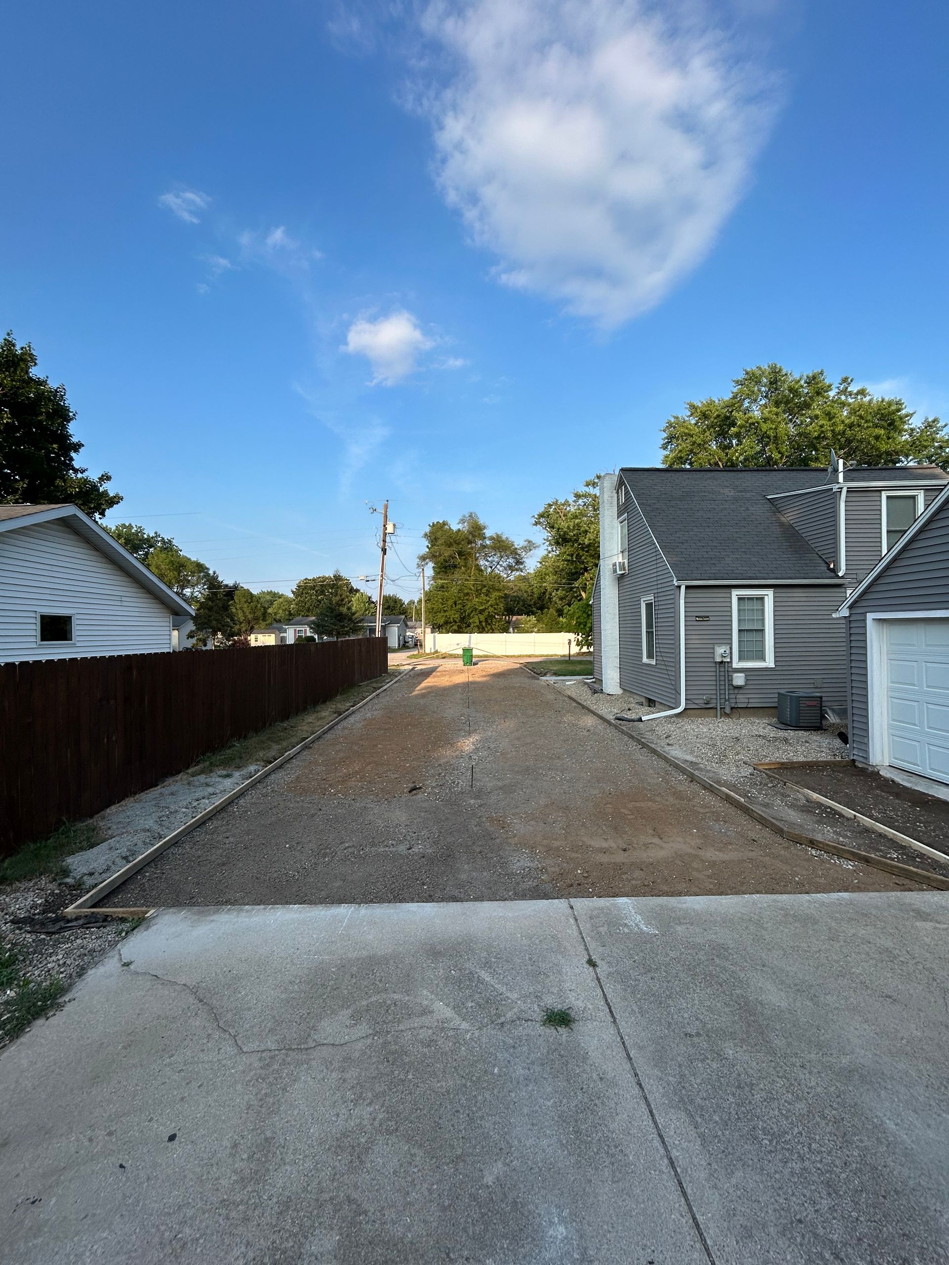 Gravel driveway between houses and a fence, under a blue sky with clouds.
