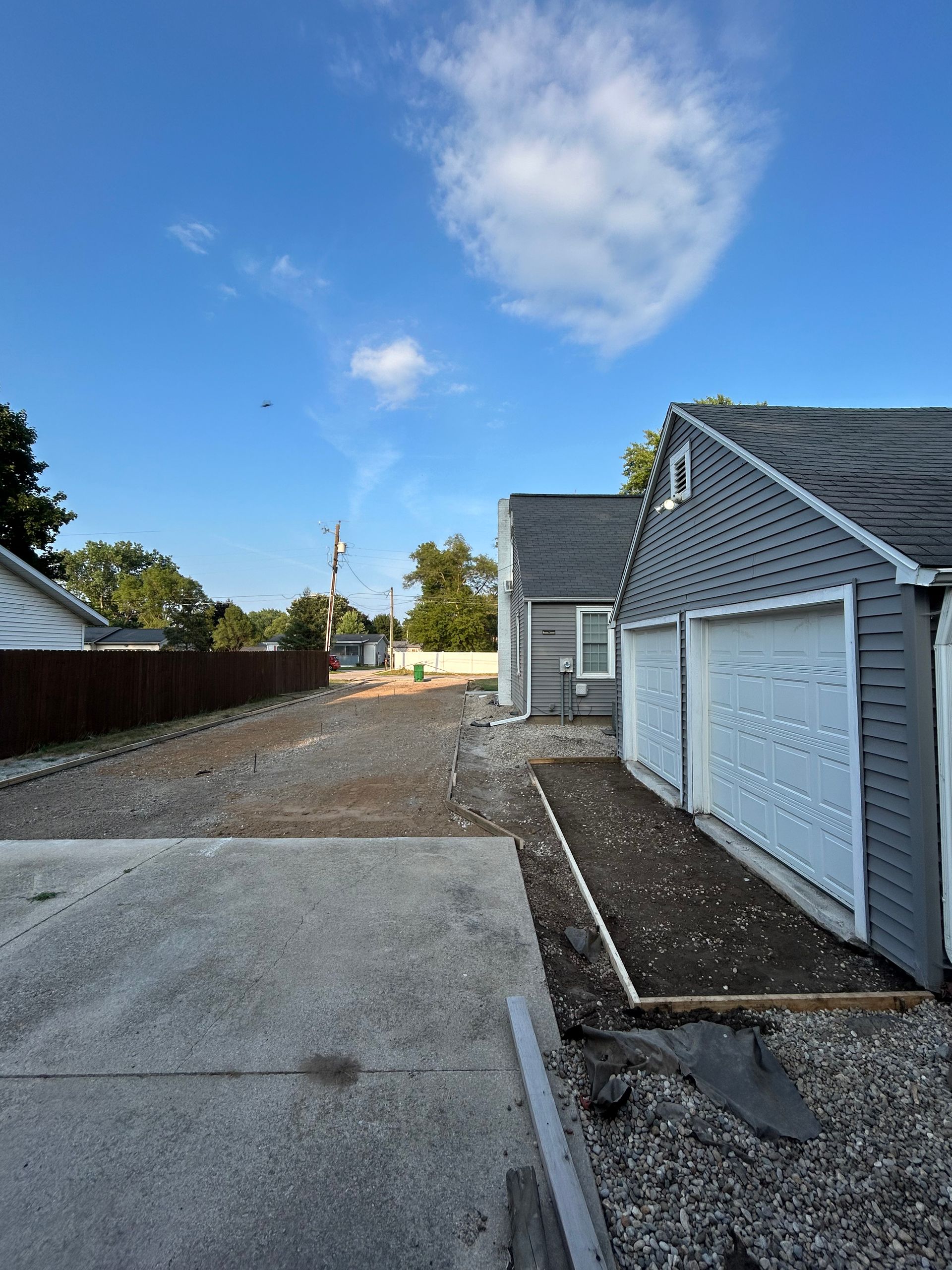 Gravel driveway with gray garage. Blue sky with fluffy clouds. Small plot of dirt.