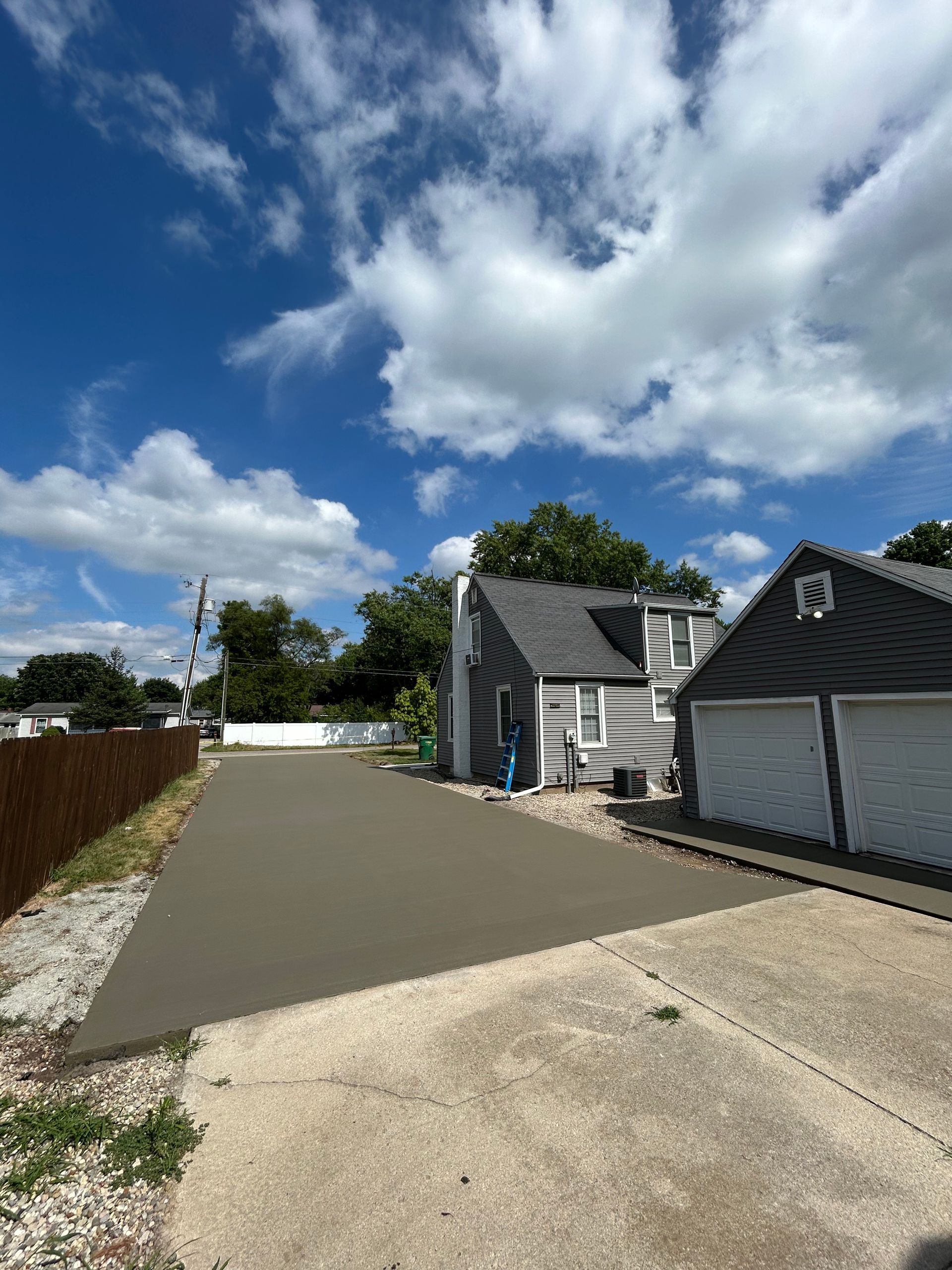 Newly poured concrete driveway next to a gray house and garage under a cloudy blue sky.