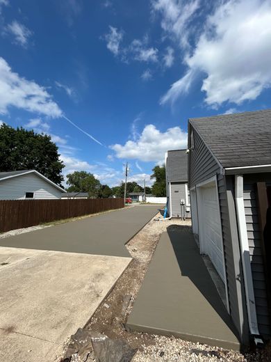 New concrete driveway being poured next to a gray garage under a blue sky with fluffy clouds.