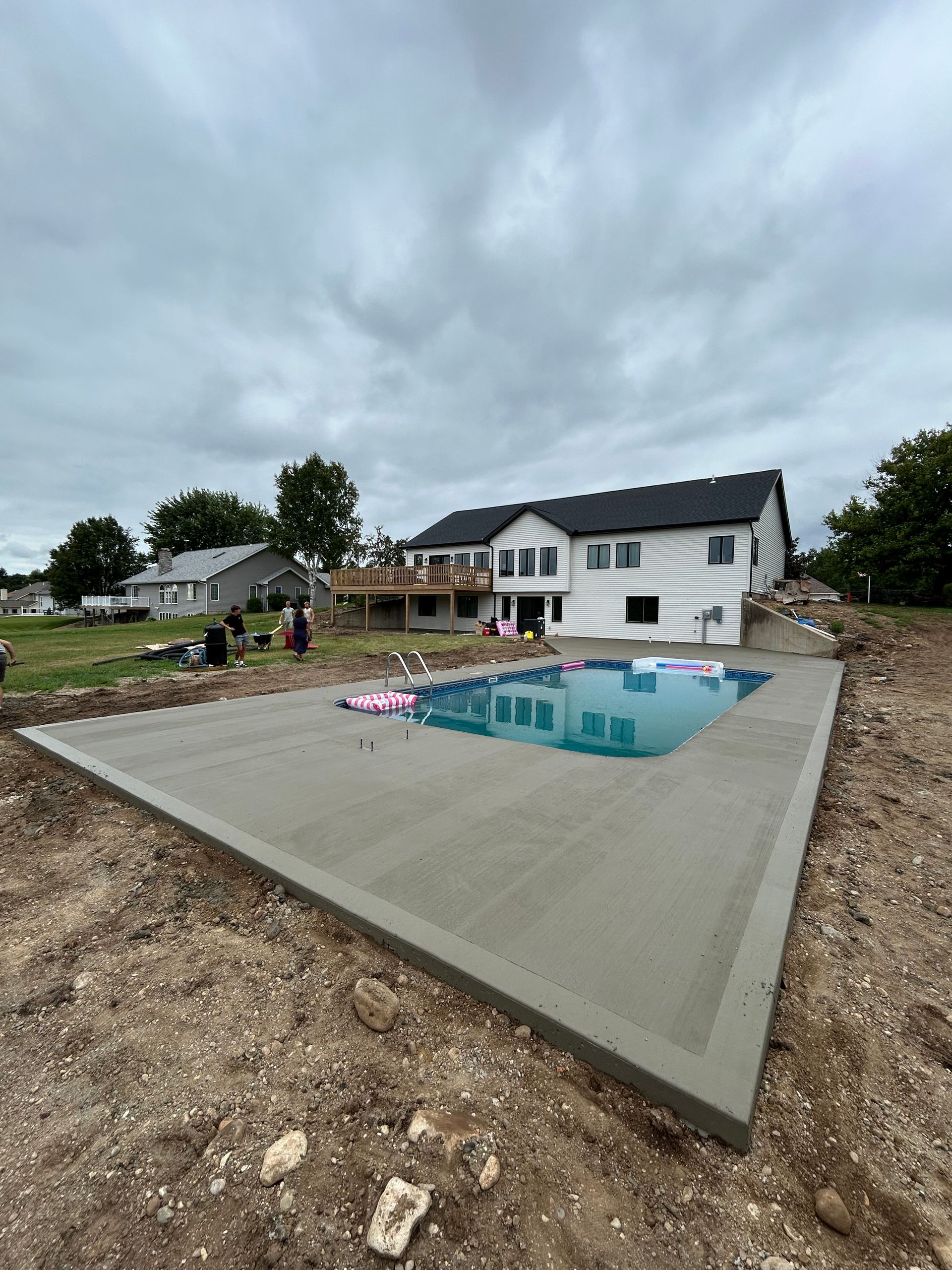 Concrete pool deck and rectangular pool next to a white house under a cloudy sky.