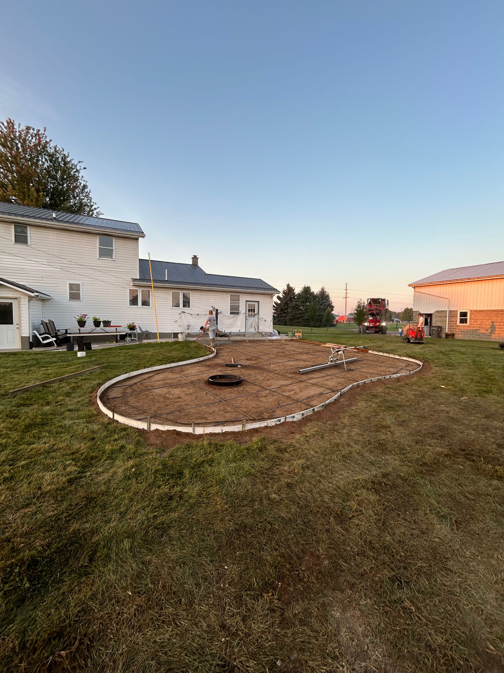 A yard with a flower bed edged with stones near a white house and a barn, at sunset.