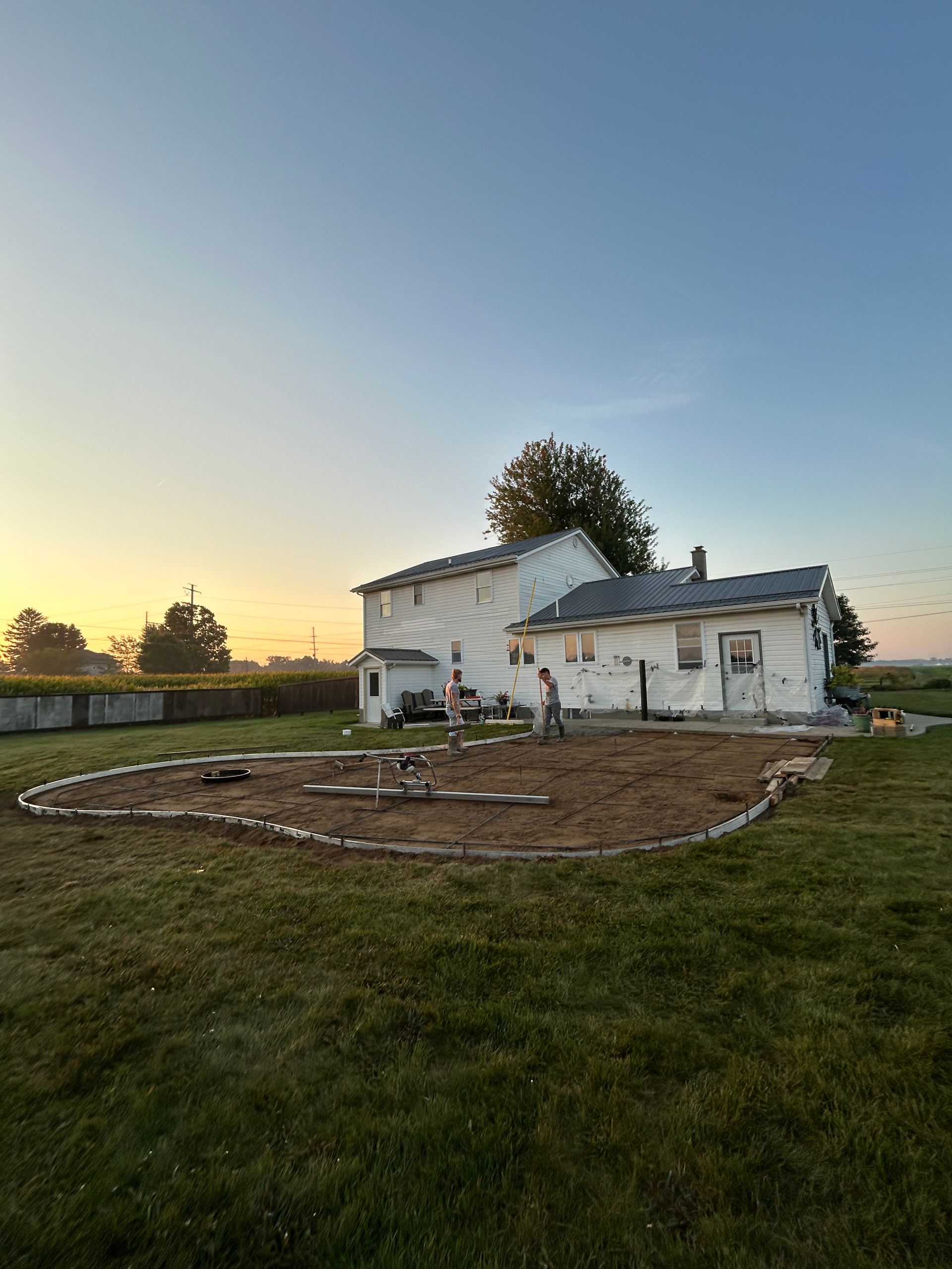 Person working in a large, dirt garden outlined with rocks next to a white house at sunset.