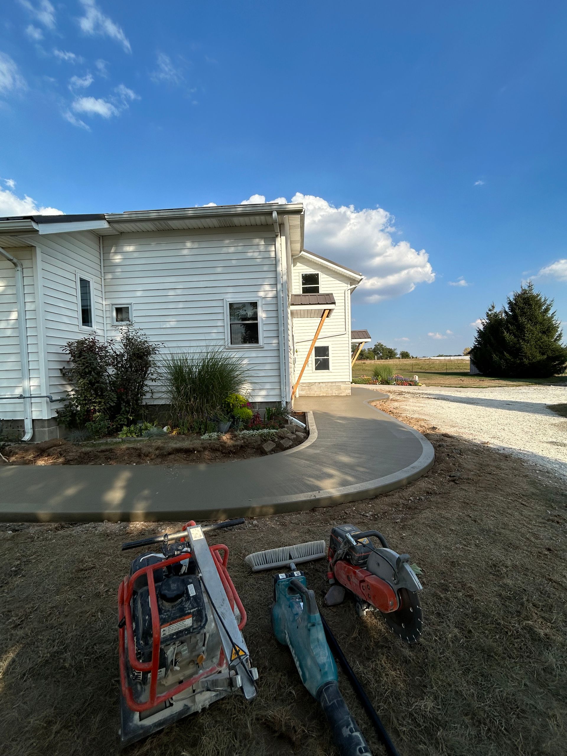 A freshly poured concrete sidewalk curves around a white building, with tools in the foreground on gravel.