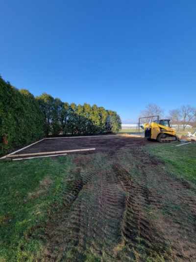 A yellow skid steer works on a cleared dirt area, framed by wooden planks, with evergreen trees and grass under a blue sky.