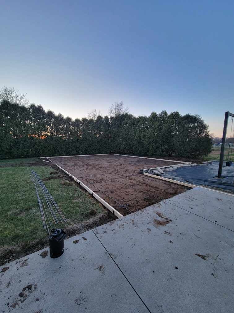 A rectangular area of dirt framed with wood next to a concrete patio, with a backdrop of trees and blue sky.