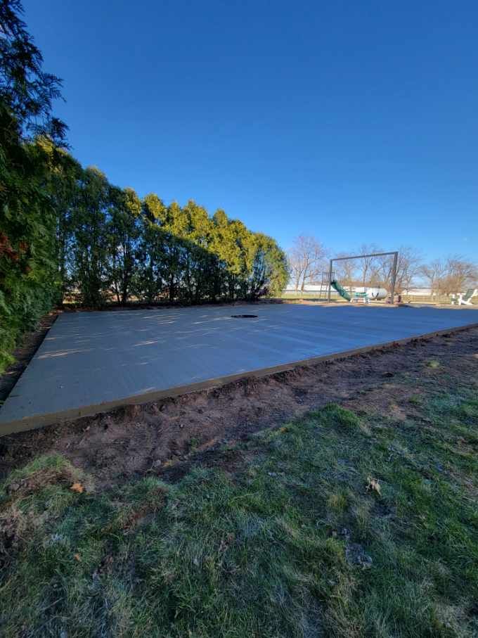 Newly poured concrete slab in a backyard, bordered by soil and grass. A swing set is visible in the distance.