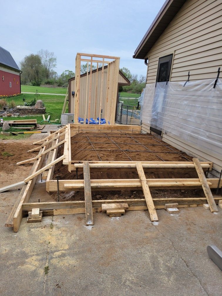 Construction site: wooden forms for a concrete patio next to a house. Tall wood frame stands nearby.