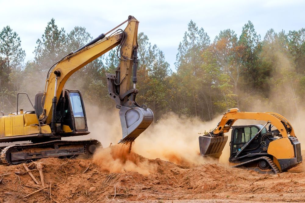 Two yellow excavators digging in dirt, creating a dust cloud in a construction site with trees in the background.