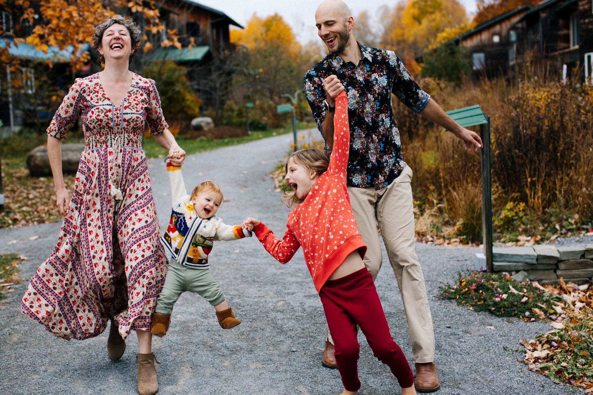Family running, holding hands, laughing on a path with fall foliage. Buildings in background.