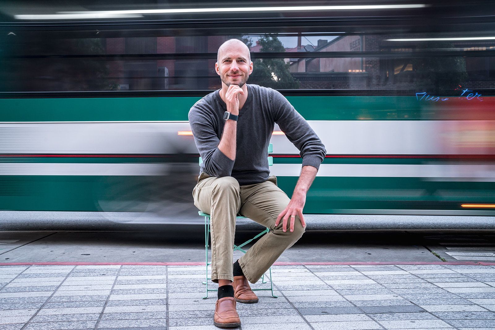 Man sits on a stool near a blurred green and white transit vehicle. He wears a gray sweater and khaki pants.