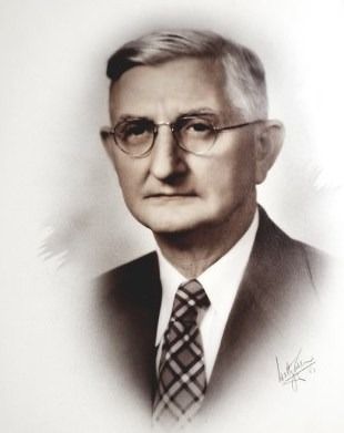 Man in glasses and suit, looking forward; sepia-toned studio portrait.