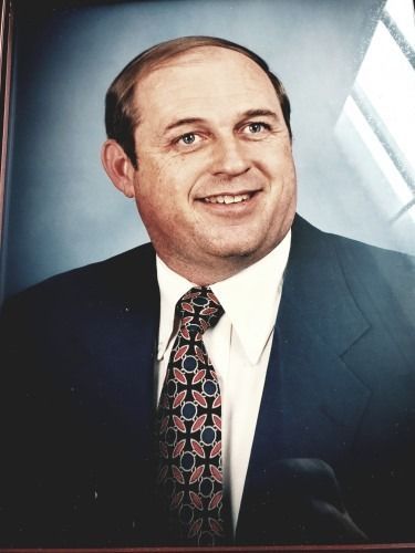 Man in a suit smiles, dark tie with red and blue pattern, against a blue backdrop.