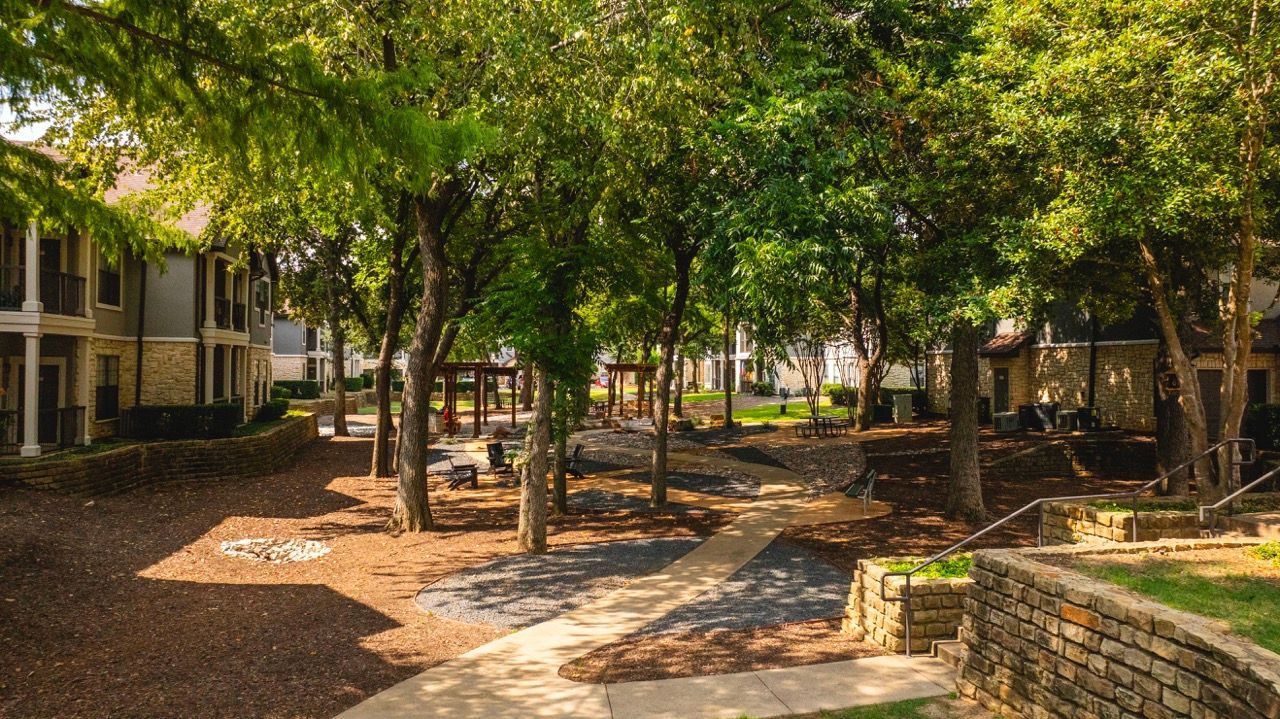 Shaded community courtyard with trees, benches, and winding walkways between apartment buildings.