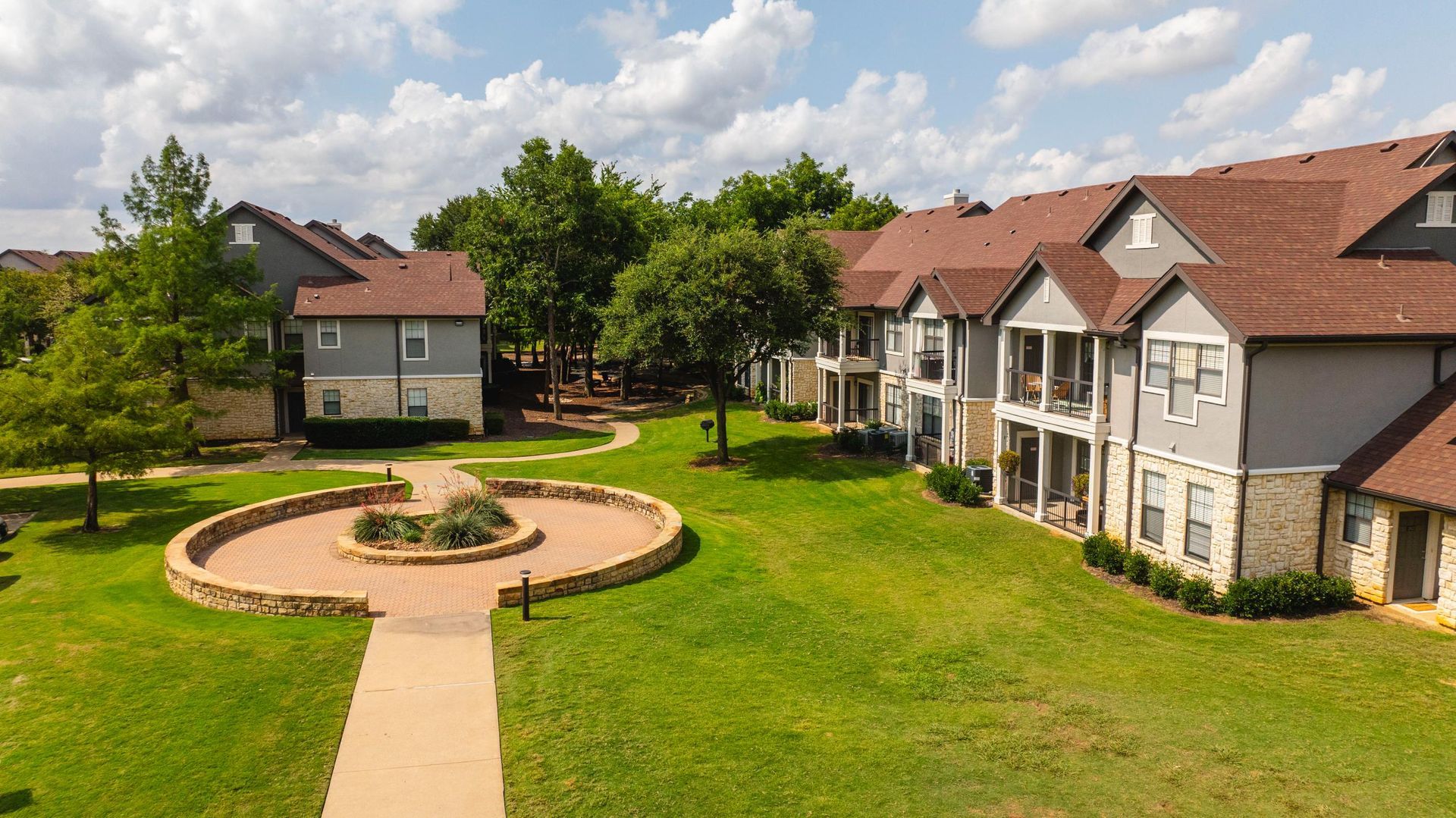 Courtyard of tan apartment buildings with red roofs, green lawn, and a circular brick planter under a partly cloudy sky
