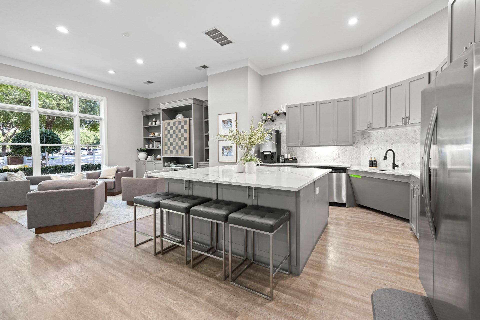 Bright modern kitchen and living area with gray island, bar stools, stainless appliances, and large windows