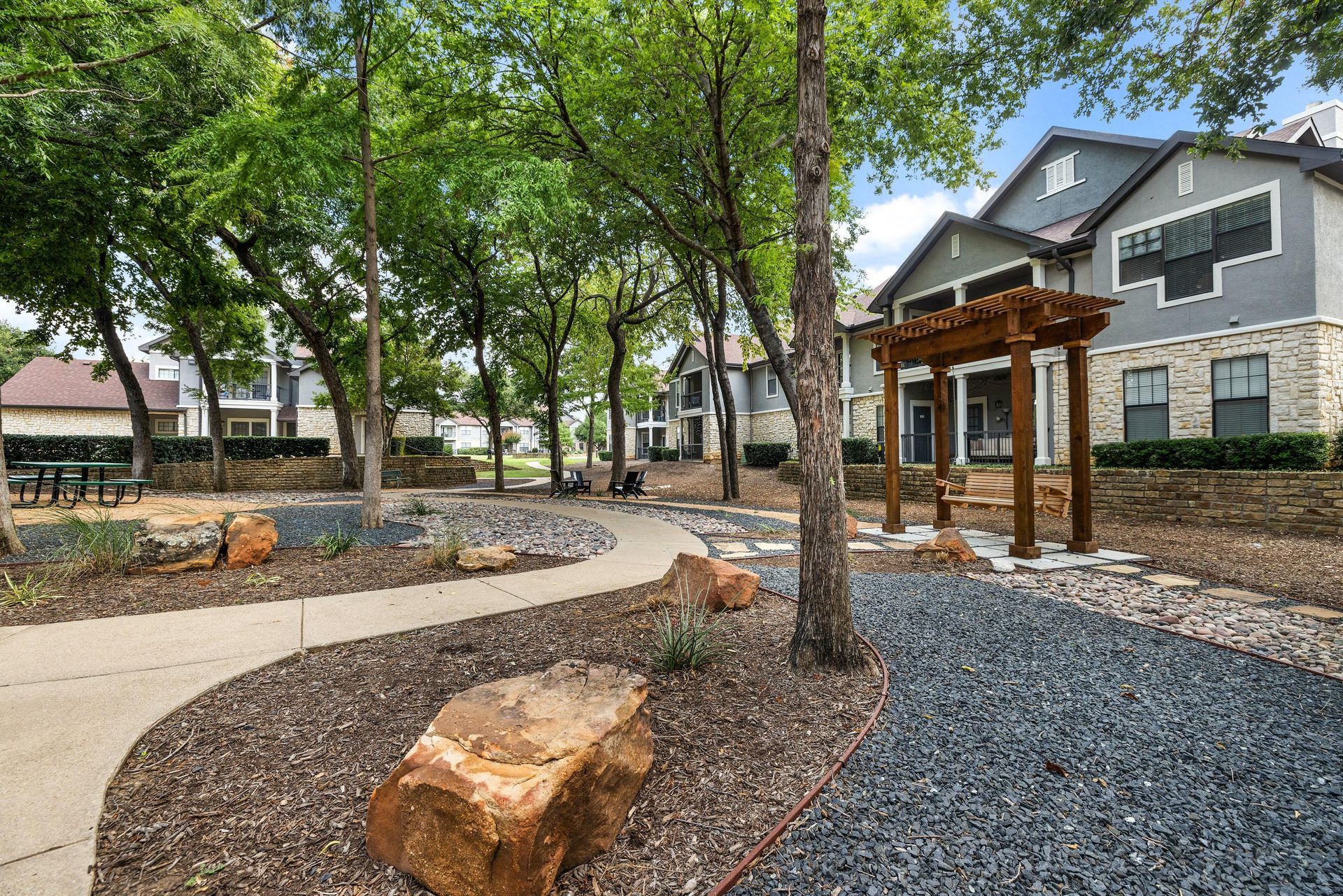 Landscaped apartment courtyard with trees, stone paths, and a wooden pergola beside brick buildings