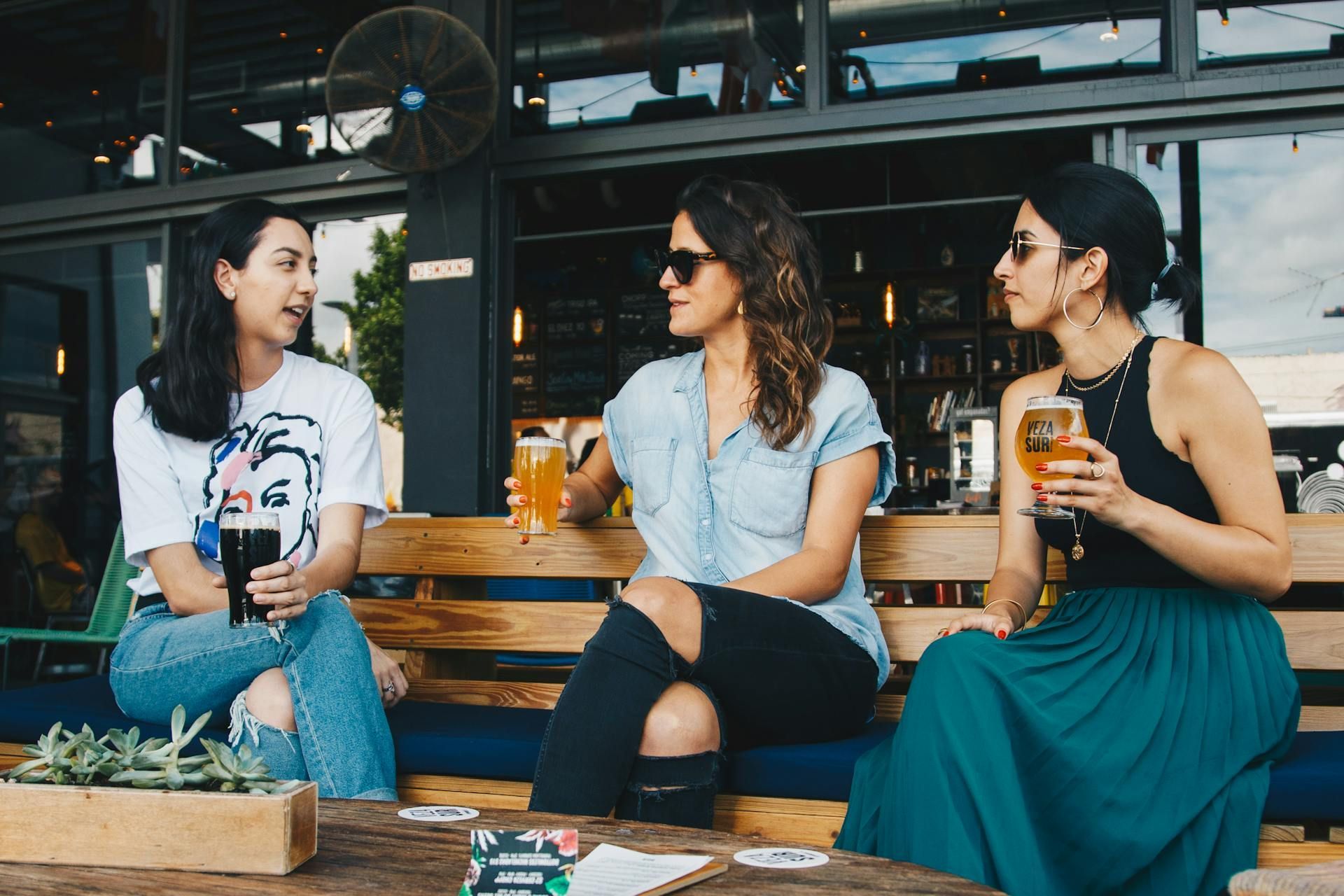 Three people sit together on an outdoor patio bench, holding drinks and talking.